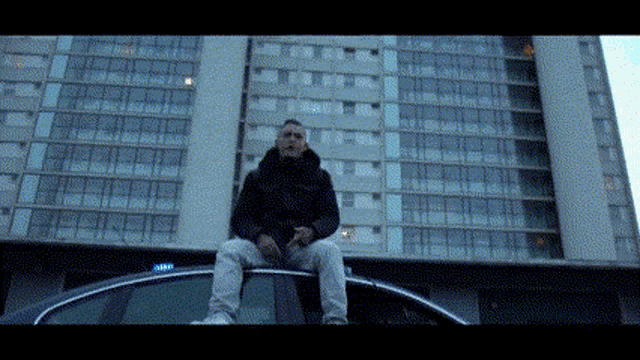 a man is sitting on the roof of a car in front of a large building .
