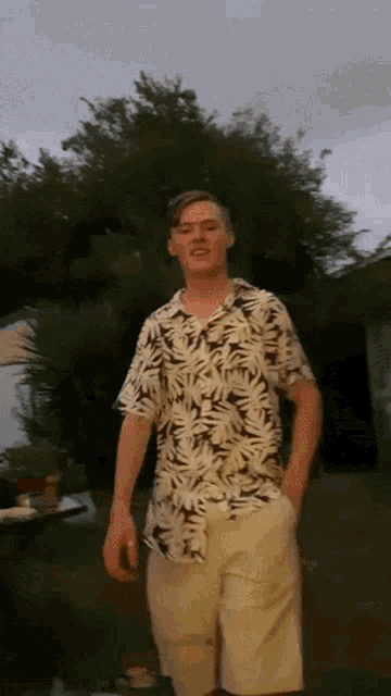 a young man in a hawaiian shirt and shorts stands in front of a tree
