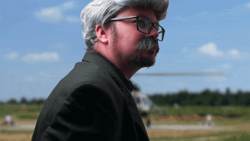 a man with glasses and a gray wig is standing in front of a helicopter