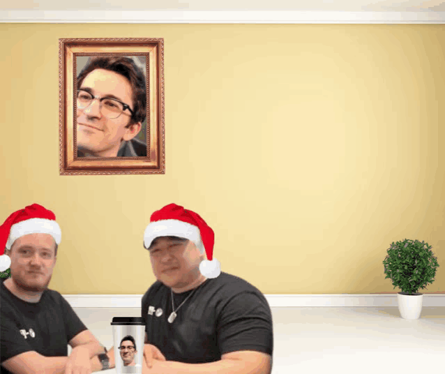 two men wearing santa hats are sitting in front of a picture of a man