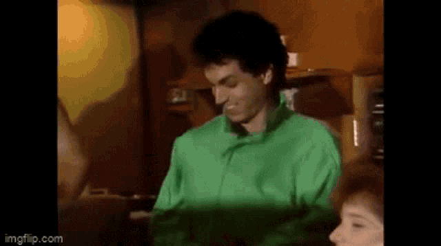 a man in a green shirt is smiling while sitting next to a woman in a room .