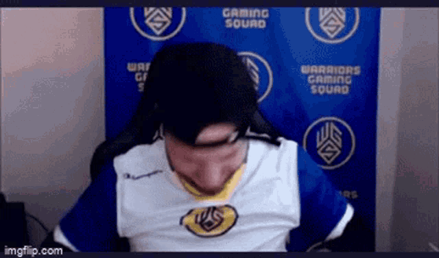 a man wearing a warriors gaming squad shirt is sitting in front of a blue wall