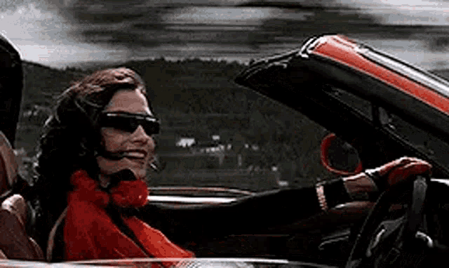 a woman wearing sunglasses and a red scarf is driving a red and black convertible .
