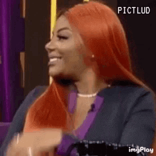 a woman with red hair is sitting in a chair and smiling while wearing a purple shirt and a black jacket .