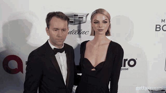a man in a tuxedo and a woman in a black dress pose on a red carpet in front of a cadillac logo