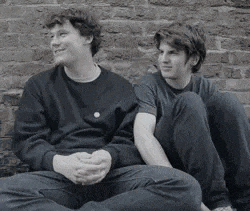 two young men are sitting next to each other on a sidewalk in front of a brick wall .