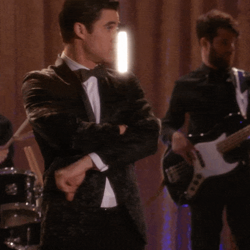 a man in a tuxedo stands with his arms crossed in front of a drum set