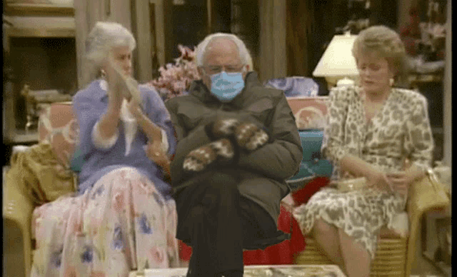 a man wearing mittens sits on a couch with two women applauding