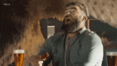 a man with a beard is sitting at a table drinking beer and eating a hot dog .