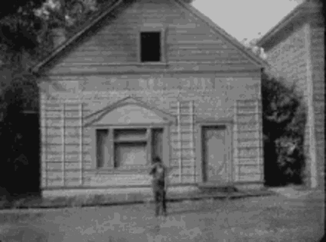 a black and white photo of a man standing in front of a small wooden building .