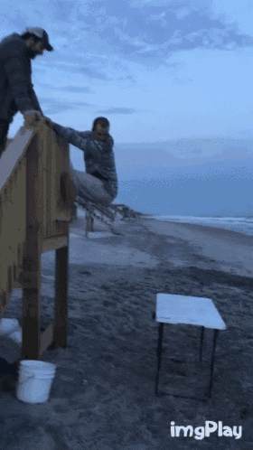 a man is sitting on a wooden structure on the beach while another man climbs on top of it