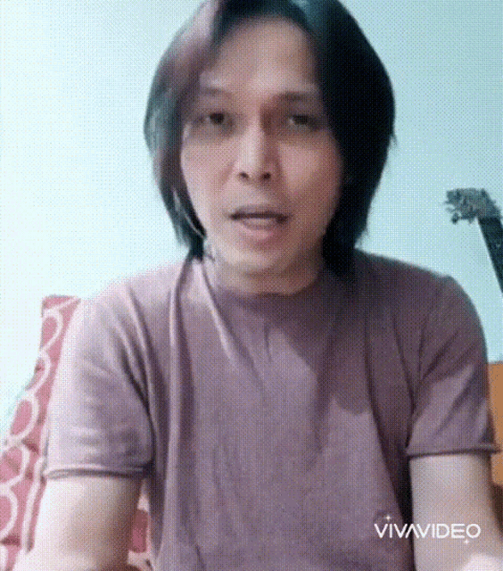 a man with long hair is wearing a purple t-shirt and talking to the camera .