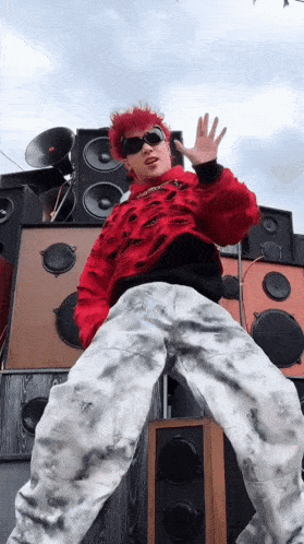a person with red hair and sunglasses is standing in front of a pile of speakers .