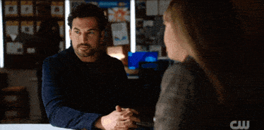 a woman in a plaid jacket is talking to a man in a dark room with a cw logo in the background