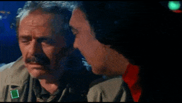 a man with a mustache is talking to another man with greek writing on the screen