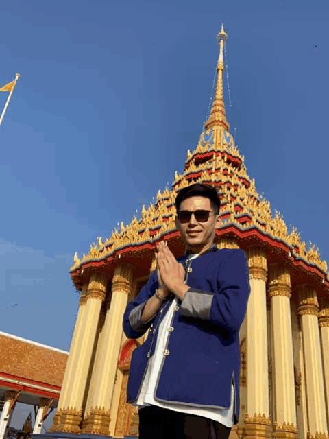 a man in a blue jacket stands in front of a temple with his hands folded