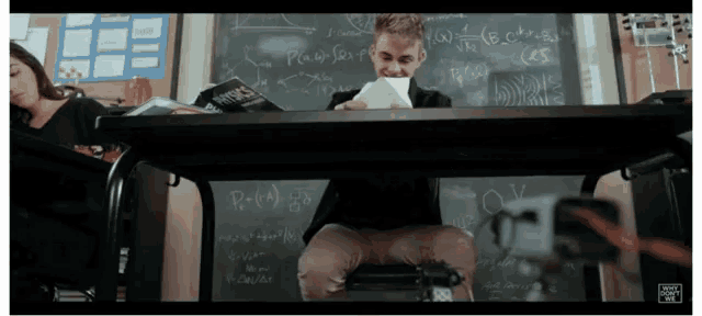 a man sits at a desk in front of a chalkboard with equations written on it
