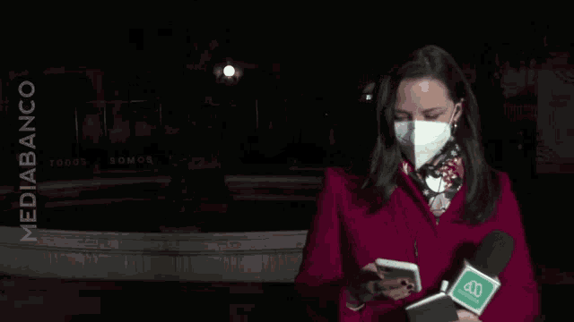 a woman wearing a mask holds a microphone in front of a sign that says todos somos todos
