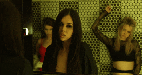 a woman looks at herself in a mirror while another woman stands in the background