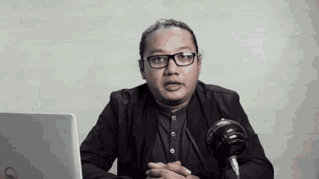 a man wearing glasses is sitting in front of a laptop and a microphone .