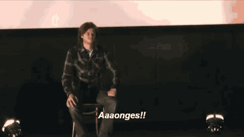 a man is sitting in a chair on a stage with the words aaaonges written on the screen behind him .