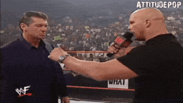 a wrestler is talking into a microphone while another man looks on .
