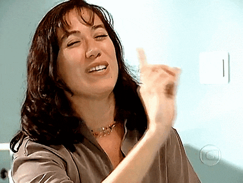 a woman wearing a necklace and a gray shirt is smiling and giving the middle finger
