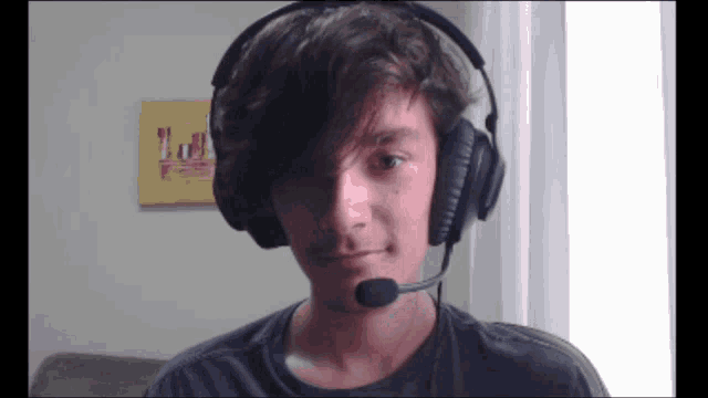 a young man wearing headphones and a microphone looks at the camera with a painting on the wall in the background