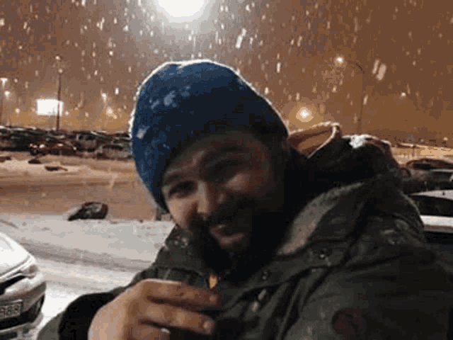 a man with a beard and a blue hat is sitting in the snow holding a cup of coffee .
