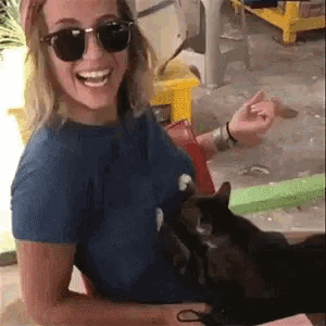 a woman wearing sunglasses is smiling while holding a black cat .