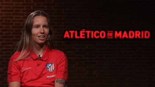 a woman in a red shirt is smiling in front of a brick wall that says atletico madrid