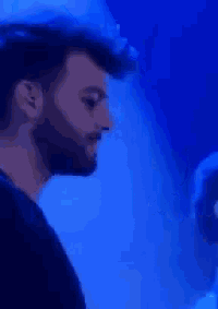 a man with a beard and a black shirt is standing in front of a blue light .