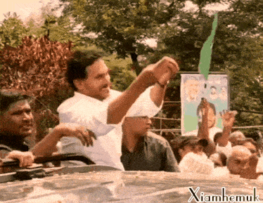 a man in a white shirt holds up a green object in front of a sign that says ' xianhemuk ' on it