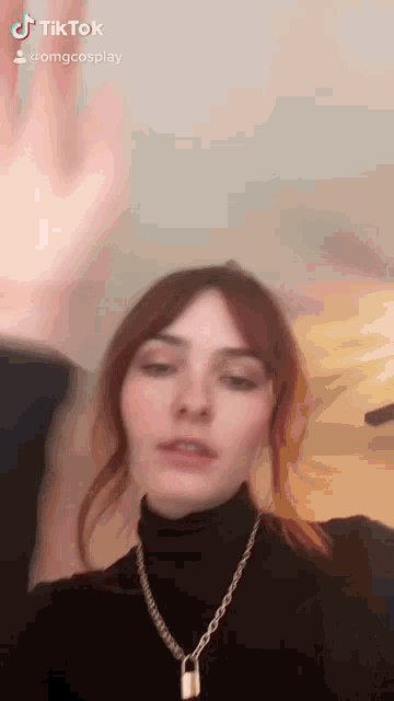 a woman wearing a black turtleneck and a necklace with a padlock on it is waving her hand .