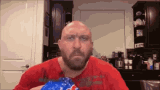 a bald man with a beard is sitting in a kitchen holding a bag of popcorn .