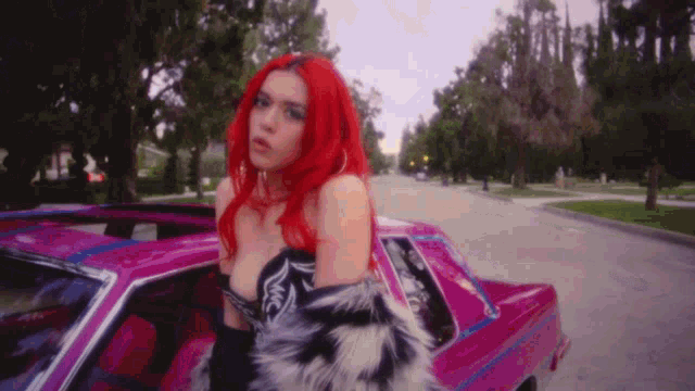a woman with red hair and a fur coat is standing next to a pink car