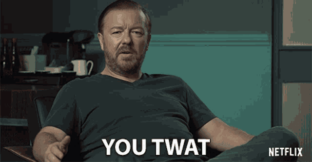a man with a beard is sitting in a chair and says " you twat "