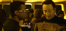 a man wearing a pair of futuristic glasses talks to another man holding a glass