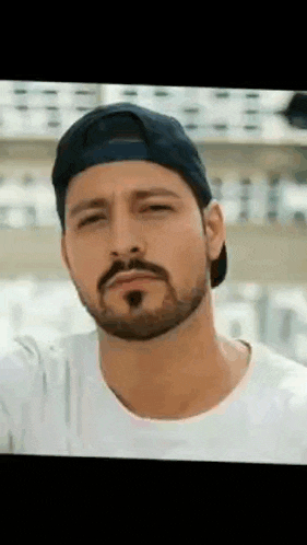 a man with a beard wearing a baseball cap and a white shirt is looking at the camera .