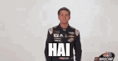 a man in a racing suit is standing in front of a white wall with the word hai on it .