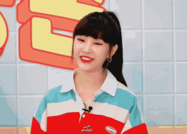 a young woman wearing a red , white and blue striped shirt is smiling while standing in front of a tiled wall .