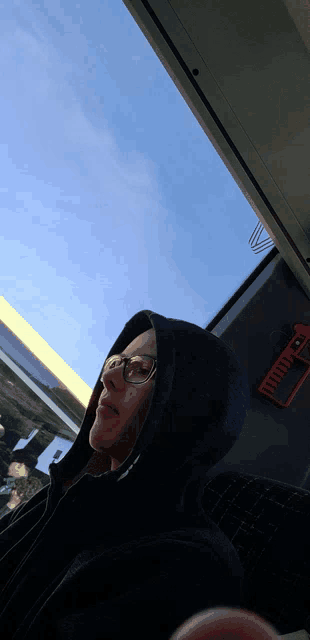 a woman wearing glasses and a hooded jacket looks out a window