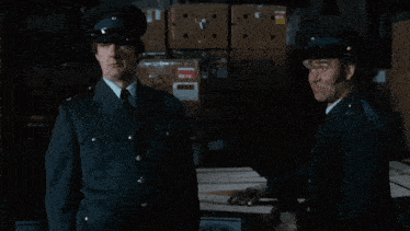 two men in military uniforms are talking in a dark room with boxes in the background