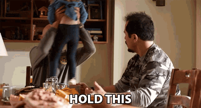 a man holds a baby in his arms while another man says hold this