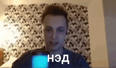 a man is holding a cell phone in front of a wall with a patterned curtain behind him
