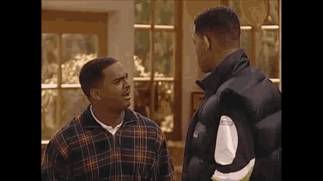 two men are standing next to each other in a living room and talking .