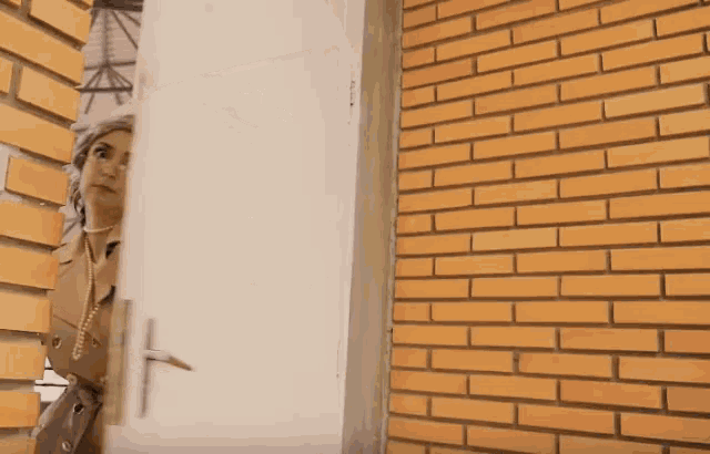a woman in a trench coat is peeking out from behind a brick wall