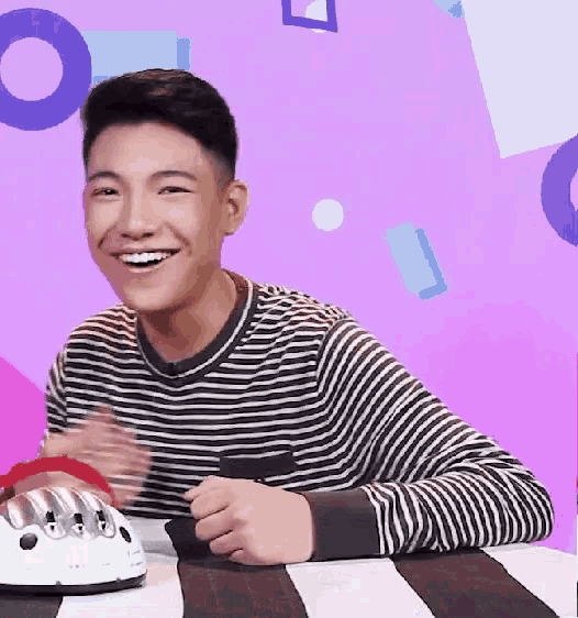a young man in a striped shirt is smiling while sitting at a table with a purple background .