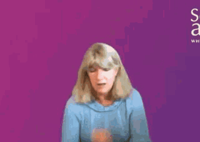 a woman in a blue sweater is sitting in front of a purple background and making a funny face .