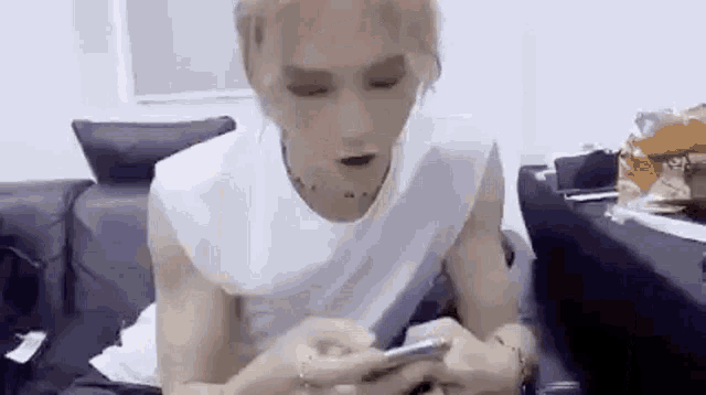 a man in a white tank top is sitting on a couch looking at his phone .